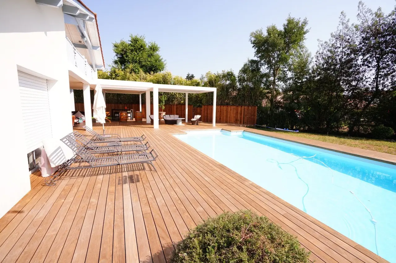 Pergola bioclimatique blanche au bord d'une piscine, terrasse en bois et transats