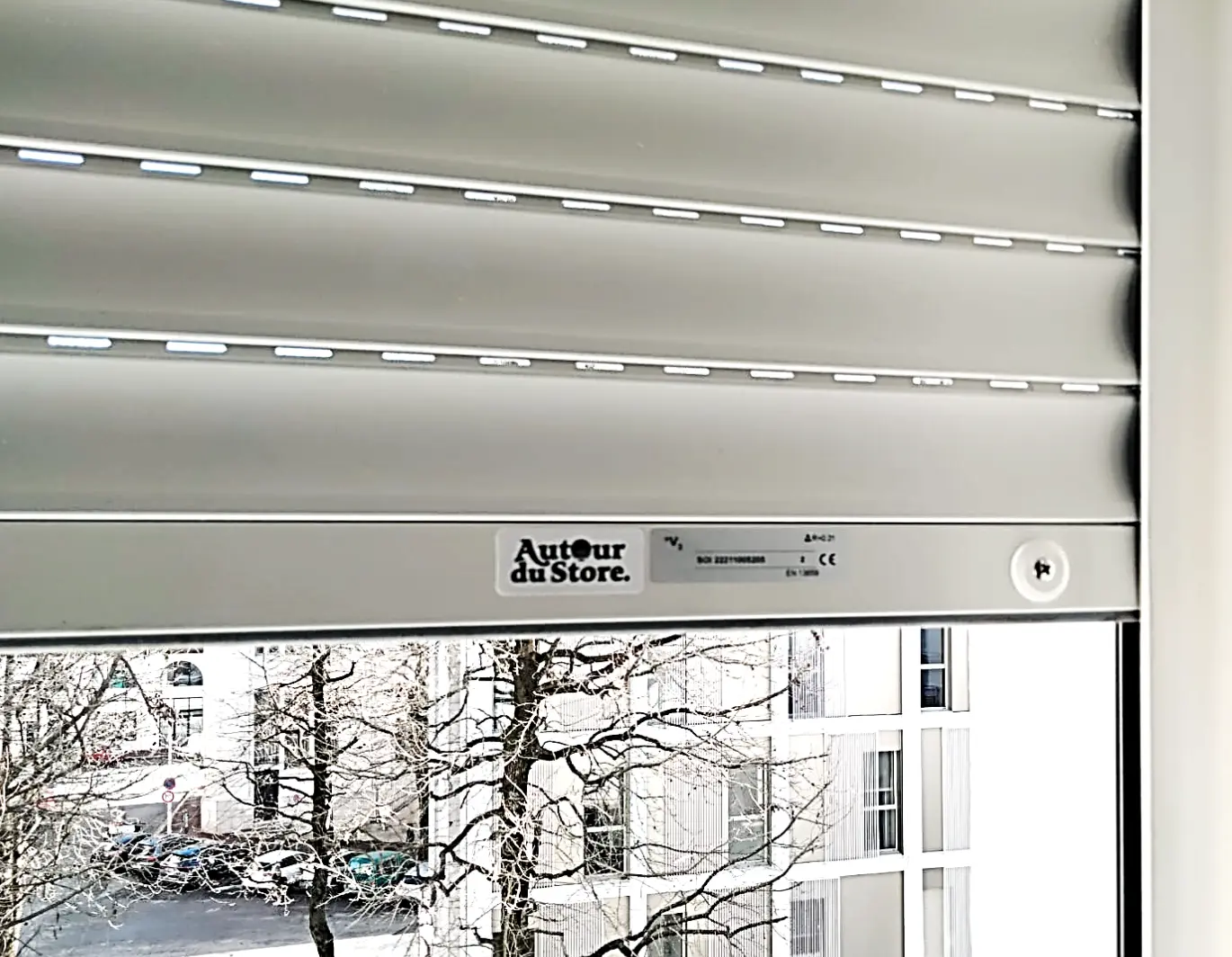 Volet roulant gris à moitié baissé, vu de l'intérieur, avec l'étiquette Autour du Store.