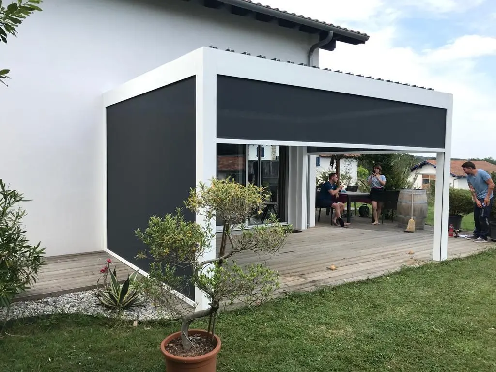 Pergola blanche adossée à une maison, côtés fermés par des stores verticaux gris