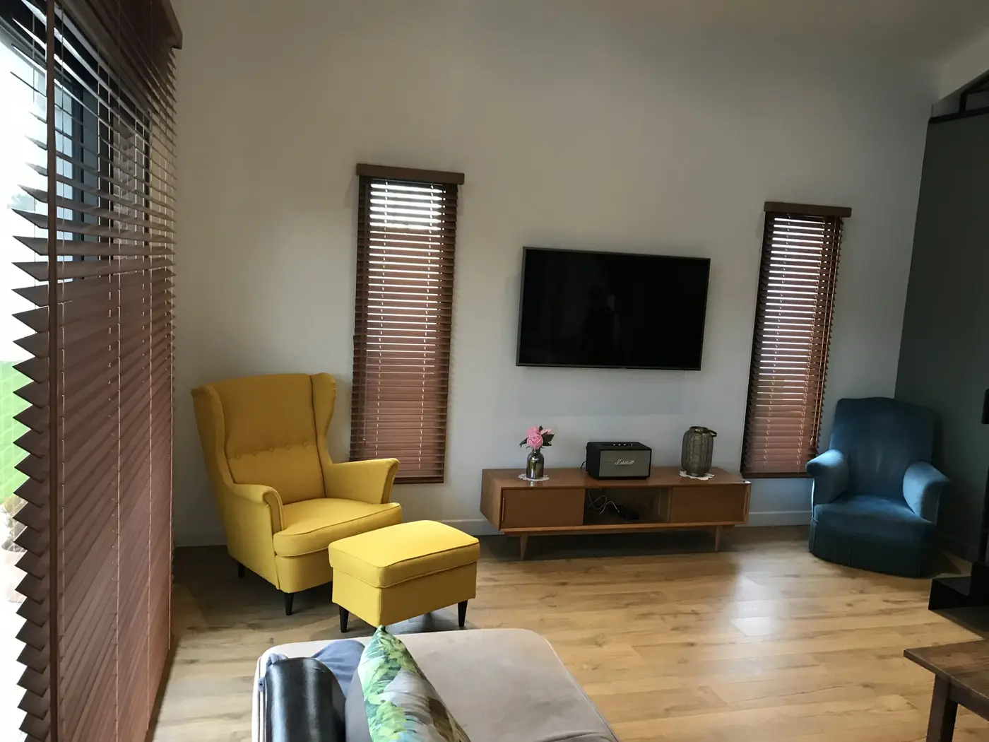 Stores vénitiens en bois sur plusieurs fenêtres d'un salon, fauteuil et pouf jaunes