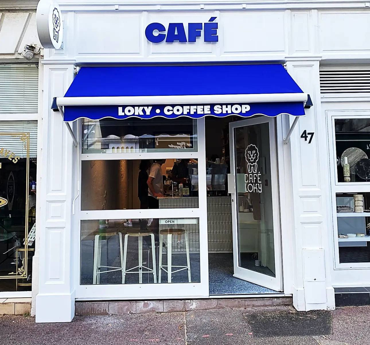 Devanture blanche d'un coffee shop avec un store bleu au-dessus de la porte vitrée.